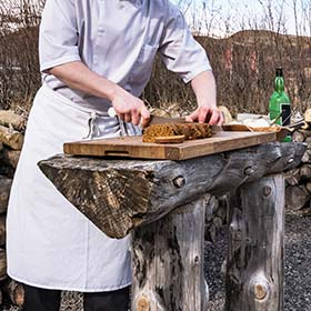 Man making traditional dark rye bread in iceland with white apron slicing bread on tree trunk